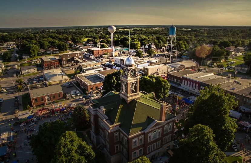 Aerial view of a small town with a historic courthouse in the foreground, surrounded by trees, and a downtown area with buildings, water towers, and people gathering on the streets. The landscape is green and stretches into the distance.